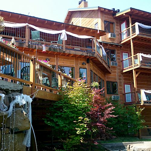 A multi-story wooden building with balconies, adorned with white fabric and plants, surrounded by greenery and rock formations, under a sunny sky.