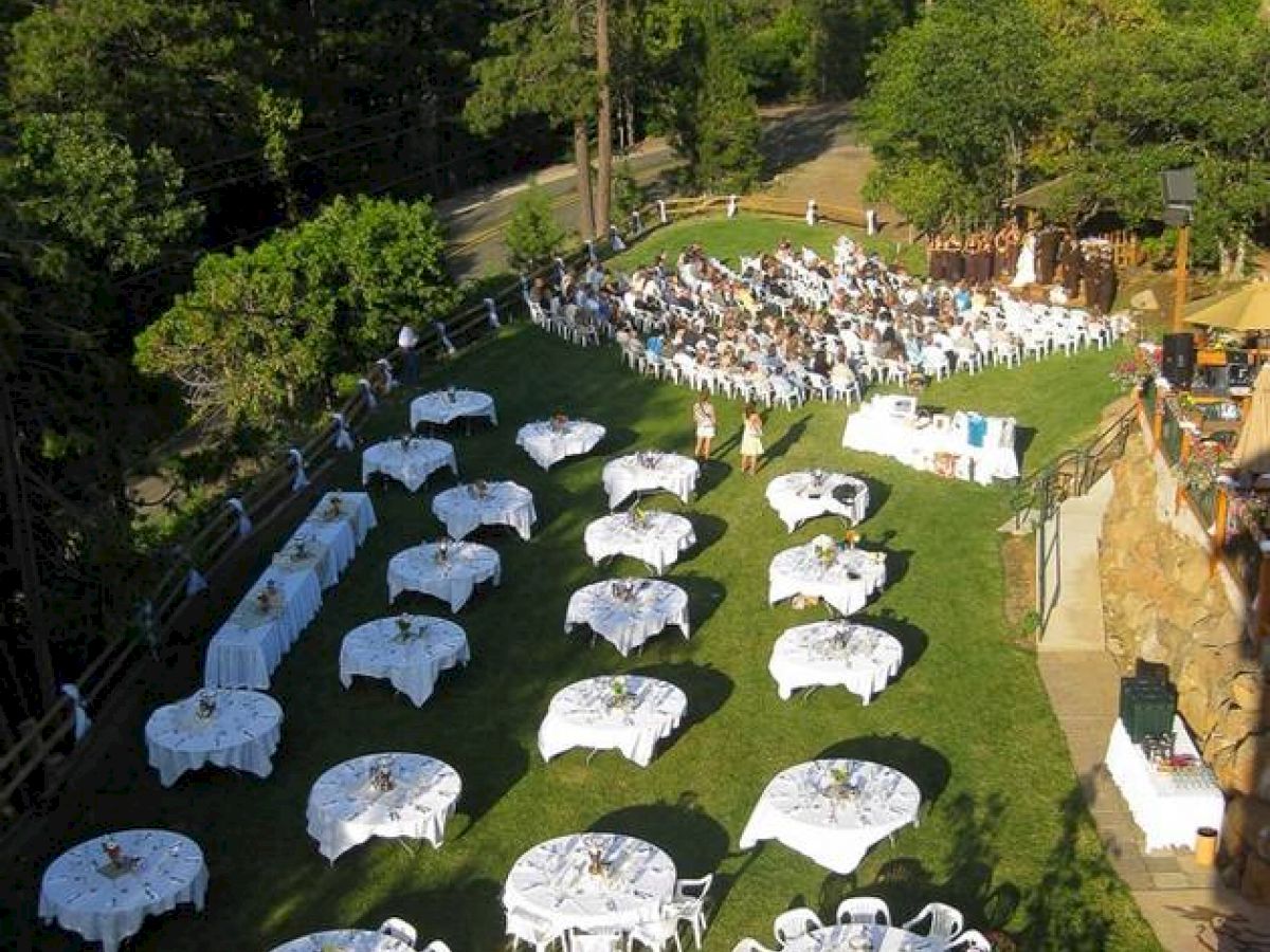 The image shows an outdoor event setup with numerous round tables covered in white tablecloths and chairs, surrounded by trees in a forested area.