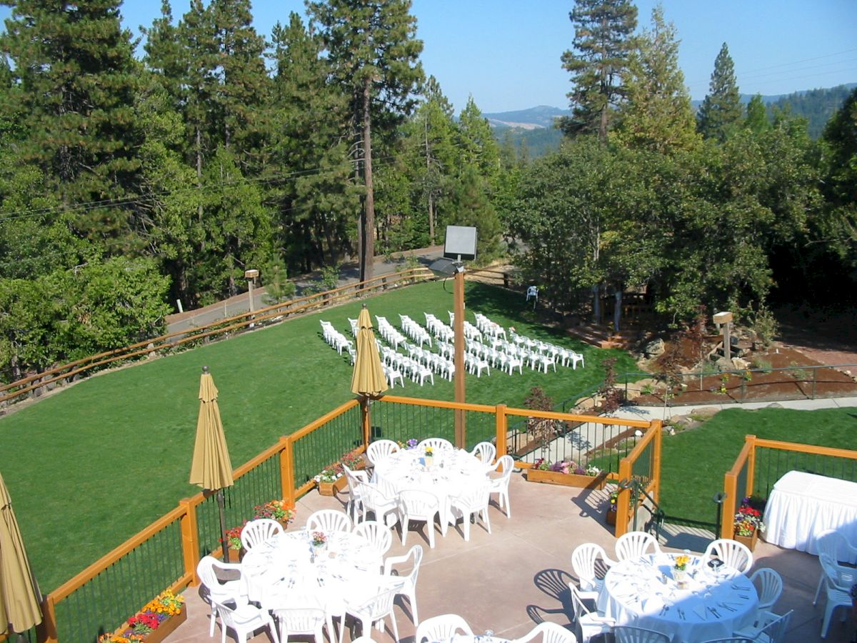 An outdoor venue with white chairs arranged on a lawn for an event, surrounded by trees, with round tables and umbrellas set up on a patio.