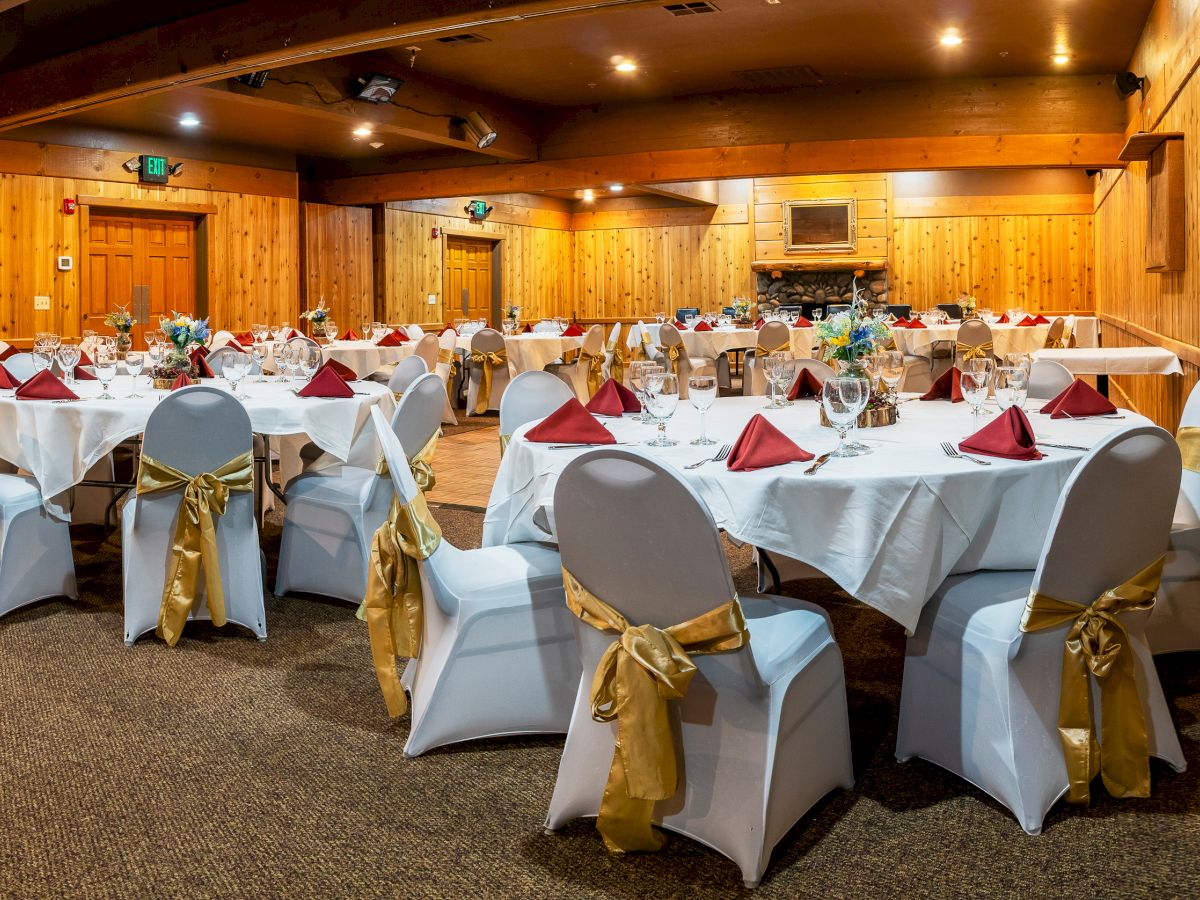 A decorated event room with round tables set for dining, white chair covers, gold sashes, red napkins, and floral centerpieces, inside a wooden venue.