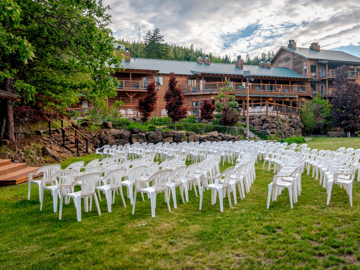 This image shows an outdoor setting with rows of white chairs facing a small wooden gazebo, and a large building in the background.