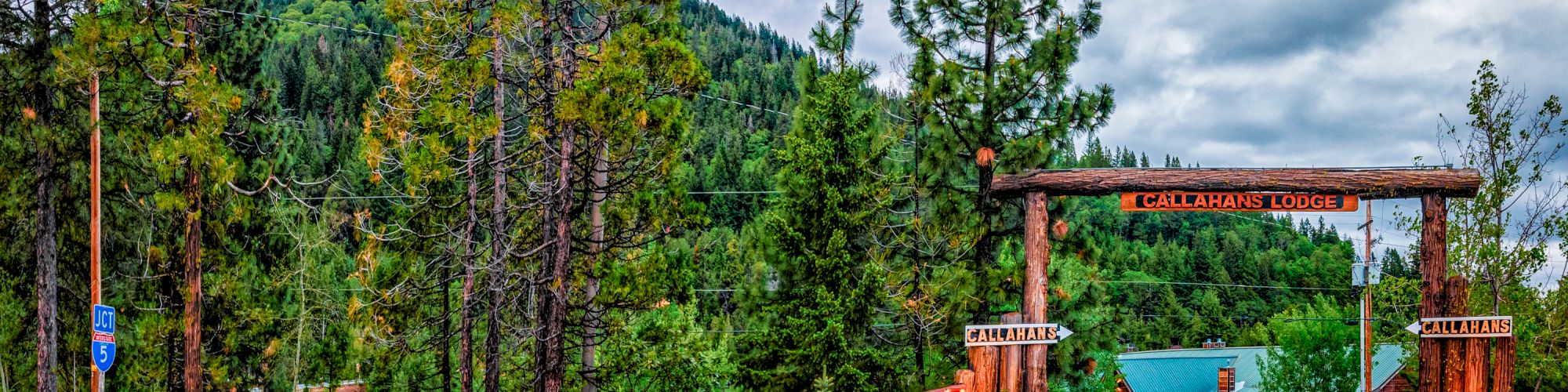 A lodge entrance amidst tall pine trees and mountains, with a wooden archway and signage, under an overcast sky.