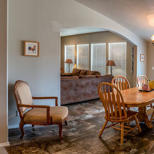 The image shows a dining area with a wooden table and chairs, a living room with a couch, lamps, and windows with blinds, and a chandelier.