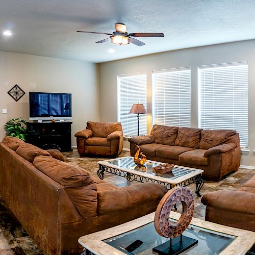 A well-furnished living room featuring brown sofas, a TV, a ceiling fan, several lamps, and decorative wall art, with a "HOME" sign on the wall.