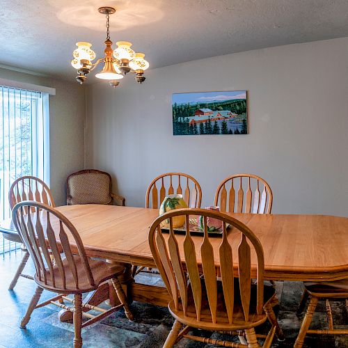 This image shows a dining room with a wooden table and six chairs, a painting on the wall, a ceiling light, and a sliding glass door with vertical blinds.