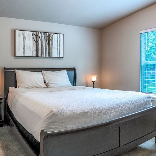 A tidy bedroom with a large bed, two nightstands with lamps, a window with blinds, and a framed tree artwork on the wall, ending the sentence.