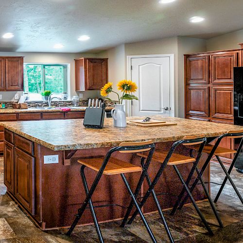 This image shows a modern kitchen with wooden cabinets, a large island with bar stools, a black refrigerator, a stove, and sunflowers as decoration.
