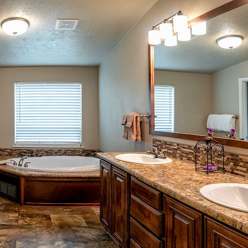 A modern bathroom featuring a double sink vanity, large mirror, bathtub with tile accents, and a window with blinds, all under warm lighting.