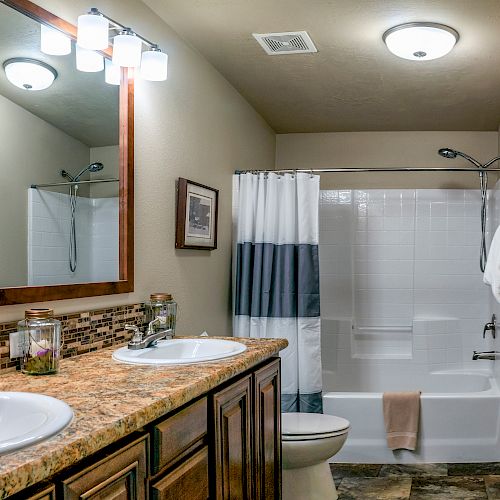 A modern bathroom with a granite countertop, double sinks, a framed mirror, a toilet, and a shower-tub combo with a striped curtain.