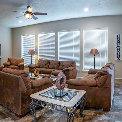 A living room with brown sofas, a TV, lamps, and decorative items on a table, with large windows and blinds, under a ceiling fan.