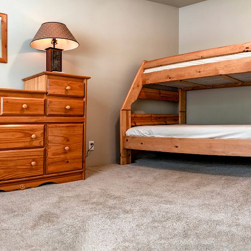 The image shows a bedroom with a wooden bunk bed, a wooden dresser with a lamp, and a small wall mirror. The carpet is light-colored.