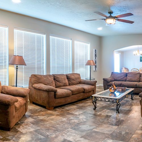 A cozy living room with brown sofas, a coffee table, lamps, and large windows with blinds. A dining area is visible through an archway in the background.
