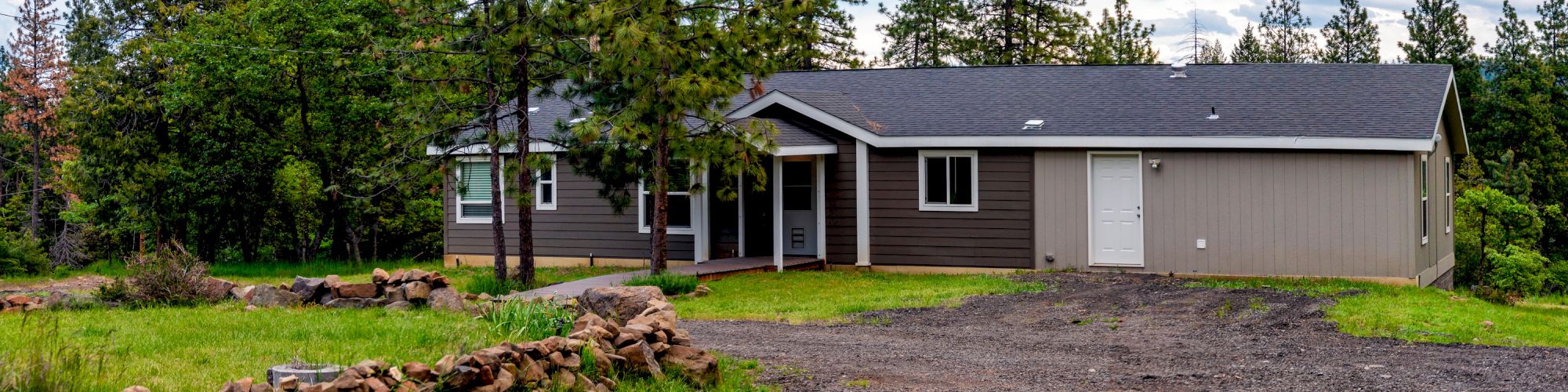 A single-story house with a gravel driveway, surrounded by trees and greenery under a partly cloudy sky.
