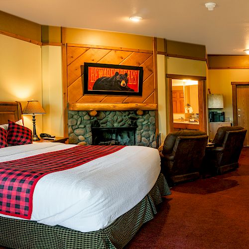 A cozy hotel room featuring a large bed with plaid decor, stone fireplace, wooden furnishings, seating area, and a small dining table.