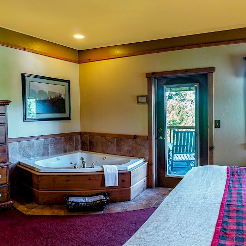 The image shows a cozy hotel room with a large bed, a jacuzzi tub, a flat-screen TV, and a dresser, leading out to a scenic balcony.