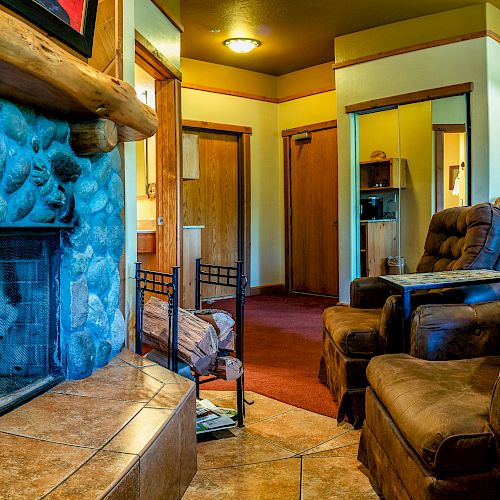 The image shows a cozy living room with a stone fireplace, two brown armchairs, and a red carpet. There are wooden doors and warm lighting.