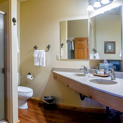 A bathroom with a large mirror, dual-sink counter, toilet, shower, and wooden accents. There are towels hanging and lights above the mirror.