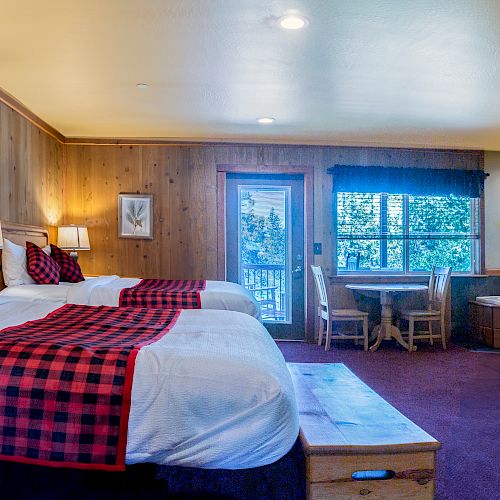 A cozy hotel room with two beds featuring red plaid linens, a wooden wall, a desk, a recliner, a TV, and a window with a view of trees.