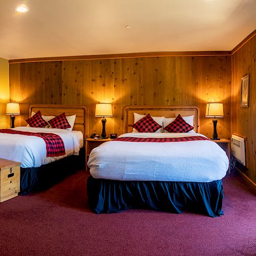 The image shows a cozy hotel room with two double beds, plaid pillows, wooden walls, lamps, and a small seating area with a table and chairs.