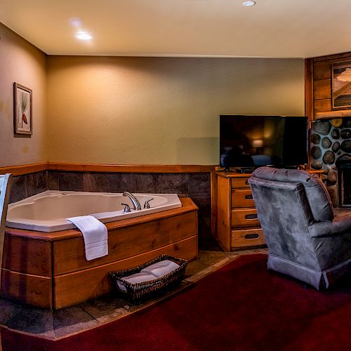 The image shows a cozy room with chairs, a round table, television, fireplace, large window, and a Jacuzzi tub next to a wood and stone accent wall.