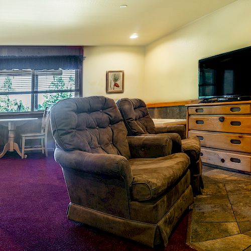 The image shows a cozy room with two plush armchairs, a flat-screen TV, and a fireplace, complemented by a wooden dresser and small table set.