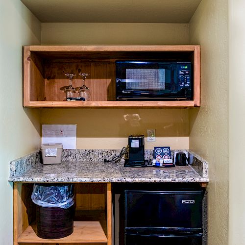 A small kitchenette with a microwave, coffee maker, mini-fridge, and shelves. There’s also a countertop with a wooden cabinet and a trash bin.