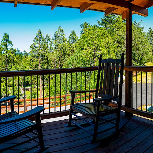 A covered porch with two rocking chairs overlooks a scenic view of trees and wooden buildings in the background, basking in bright sunlight.