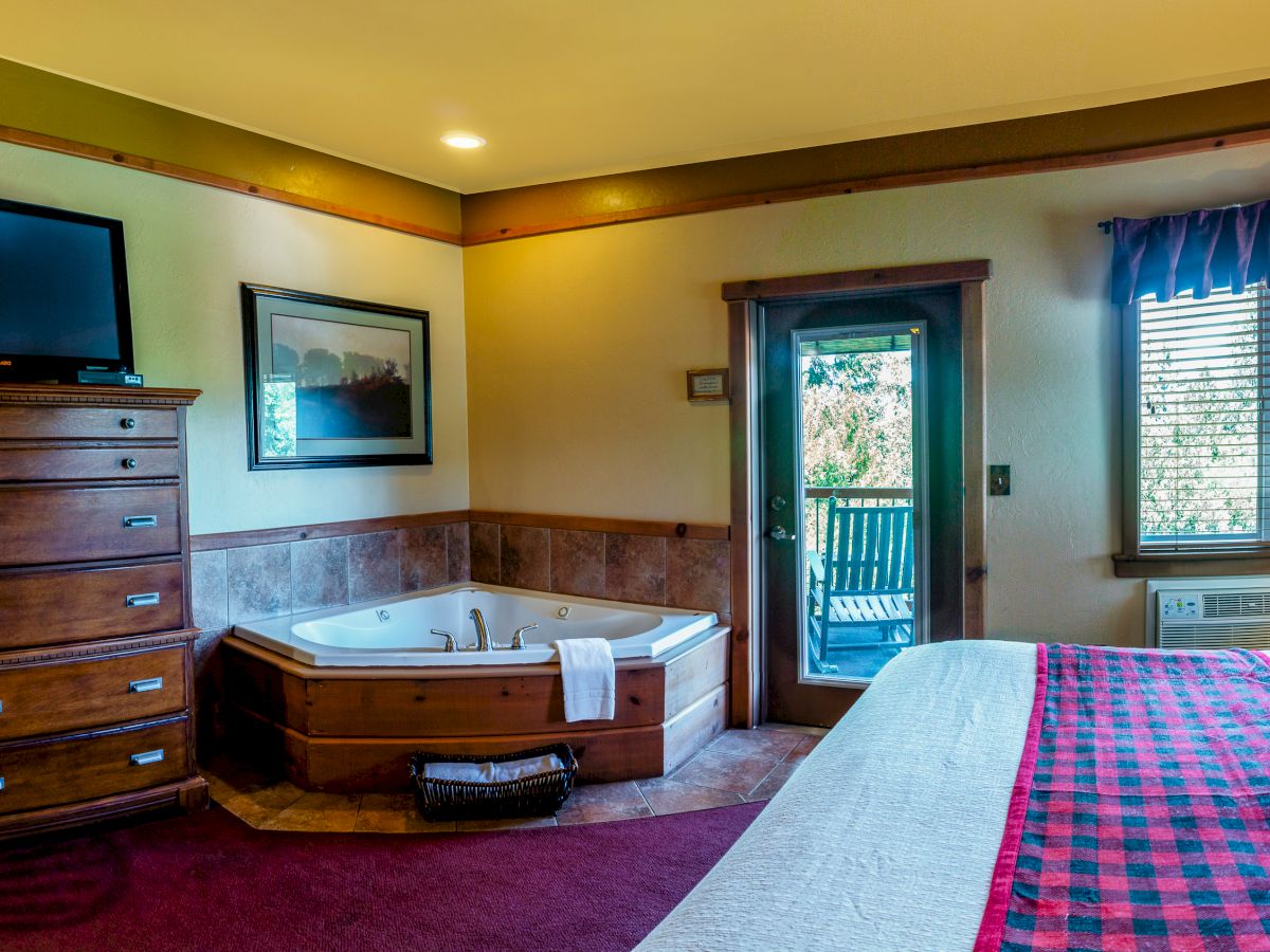 A bedroom with a bed, TV on a dresser, a jacuzzi, a door to a balcony, a window, and warm lighting.