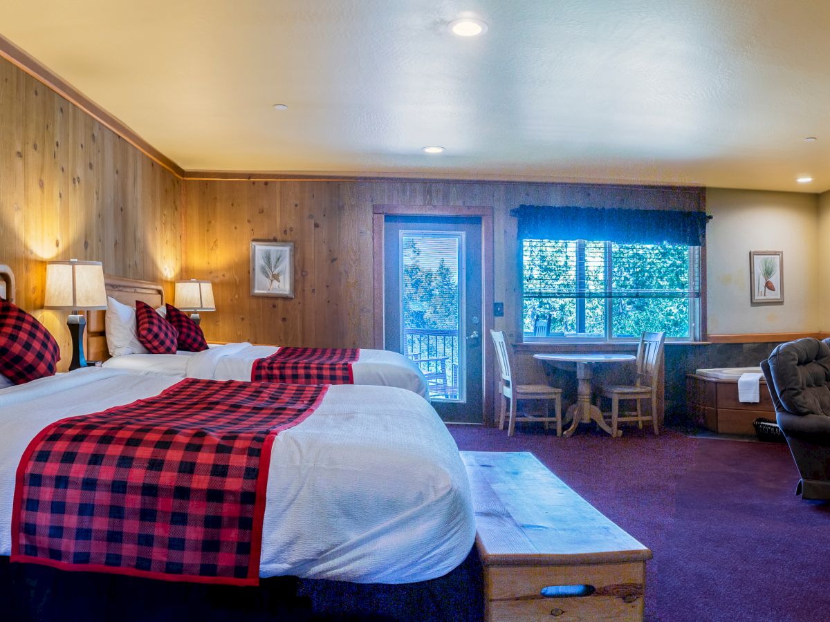A cozy room with two beds, checkered linens, a small dining area, a recliner, a TV, and wooden decor. The room has a warm, cabin-like feel.