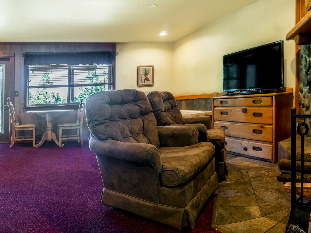 A cozy living area featuring two plush armchairs, a fireplace, a flat-screen TV, a wooden dresser, and a desk with chairs by the window.