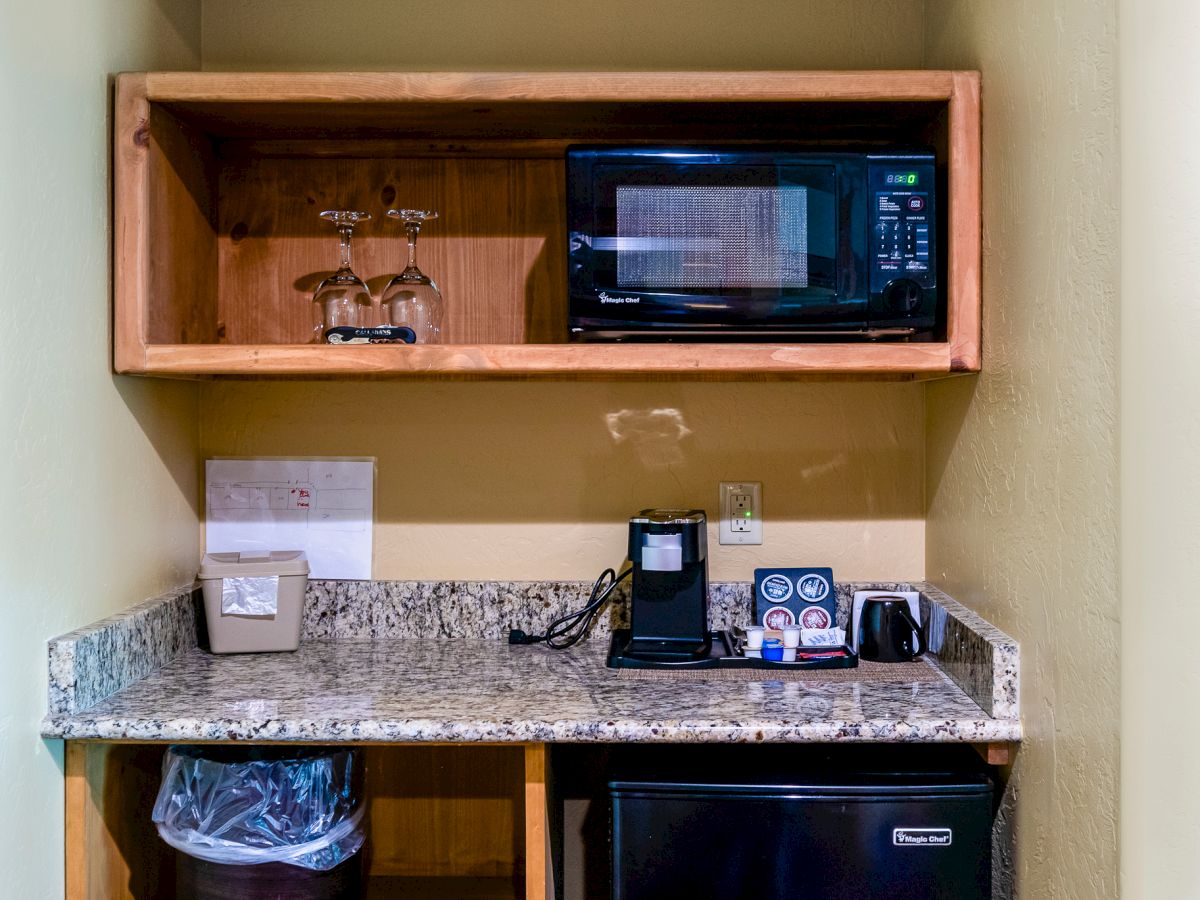 A small kitchenette with a microwave, a coffee maker, a mini-fridge, and a trash bin under a granite countertop in a wooden cabinet.