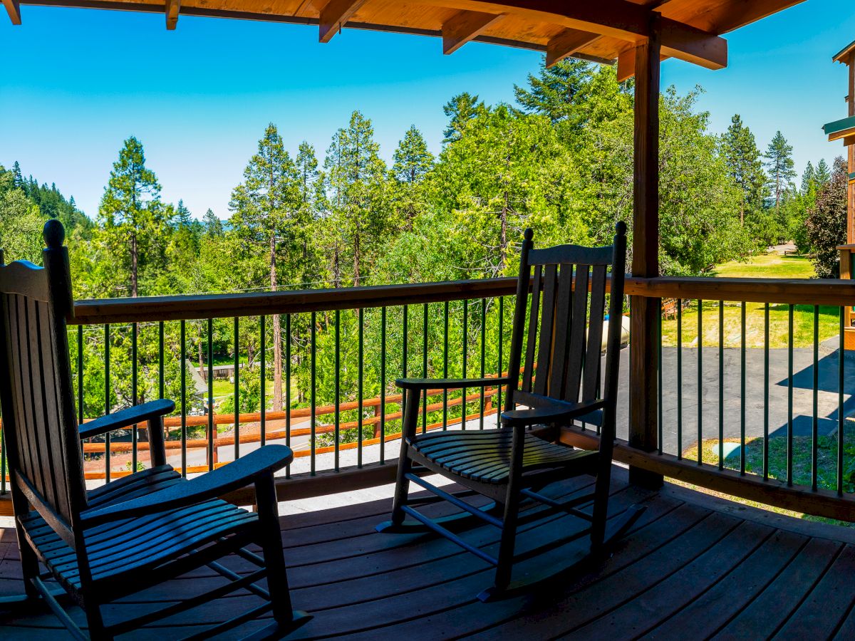 A wooden porch with two rocking chairs overlooks a scenic view of trees and wooden buildings on a bright, sunny day.