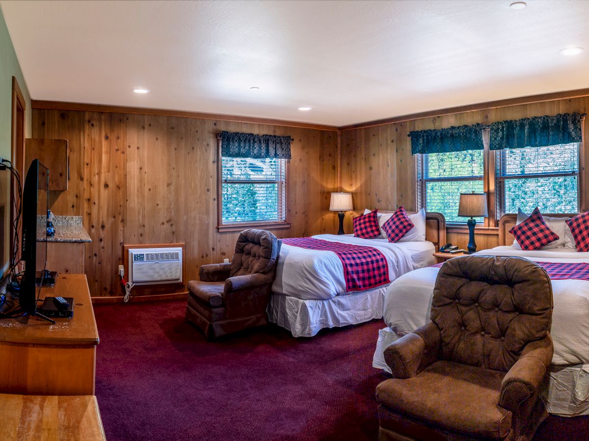 This image shows a cozy hotel room with two beds, armchairs, wooden walls, a TV, and windows with curtains. The ambiance is rustic and inviting.