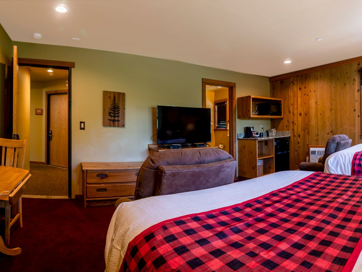 A cozy hotel room with two beds, a small table, chairs, a TV, and a kitchenette area. The decor features a mix of wooden and green elements.