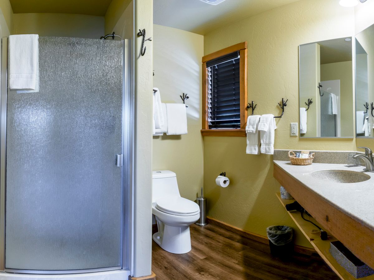 A bathroom with a shower, toilet, sink, mirror, towel racks, wooden floor, wastebasket, and window with blinds, all under bright lighting.
