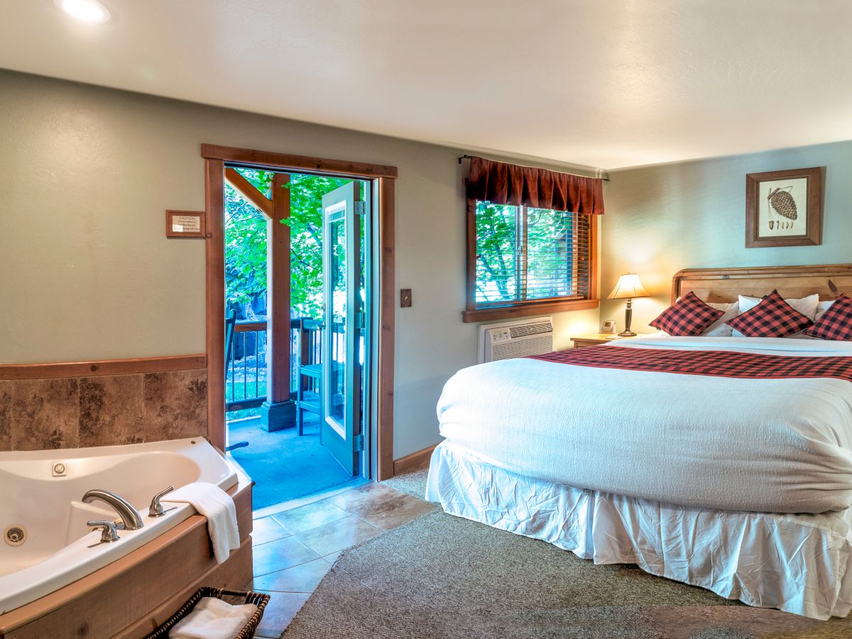 A cozy bedroom with a king-size bed, jacuzzi tub, wooden furniture, and access to a patio with a scenic outdoor view, warmly lit with lamps.