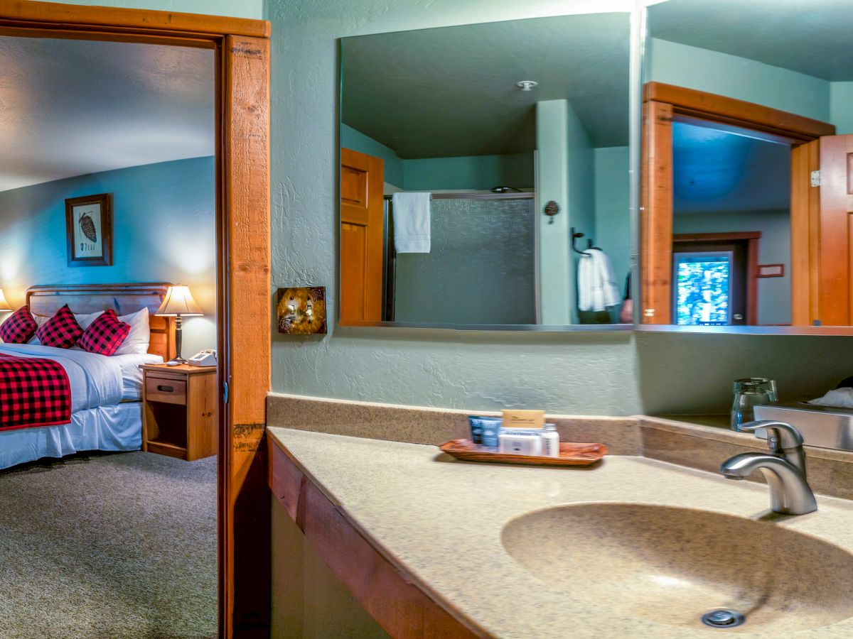 A cozy bedroom with a bed, nightstands, and a red plaid theme is visible from a bathroom with a countertop, sink, and mirrors.