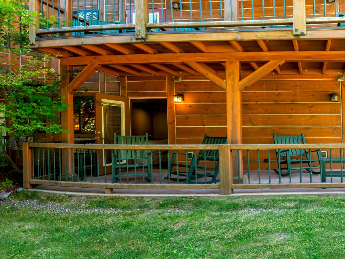 The image shows the exterior of a wooden building with a porch featuring several rocking chairs and a railing, surrounded by greenery and a grassy area.
