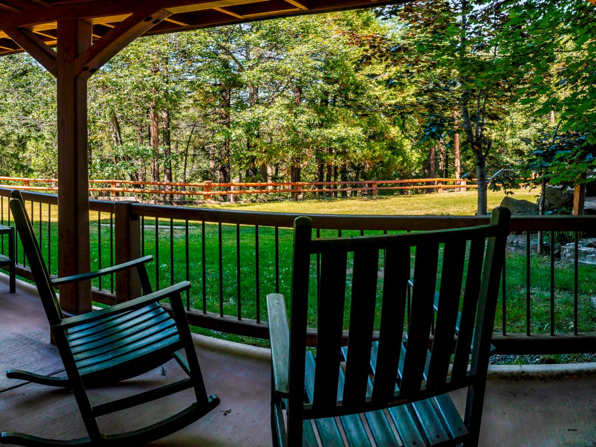 A covered porch with rocking chairs overlooks a green lawn and a wooded area in the background, creating a peaceful and natural setting.