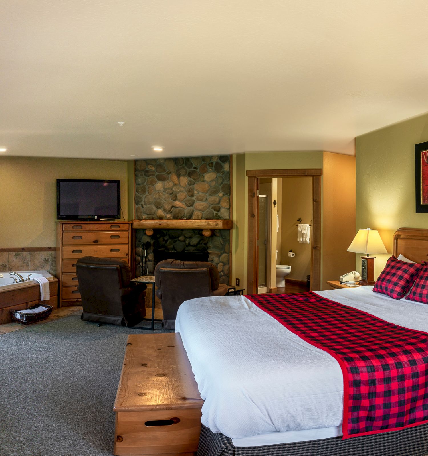 A cozy hotel room with a king-sized bed, plaid-themed decor, a fireplace, a TV, a jacuzzi, and wooden furniture for a rustic feel.