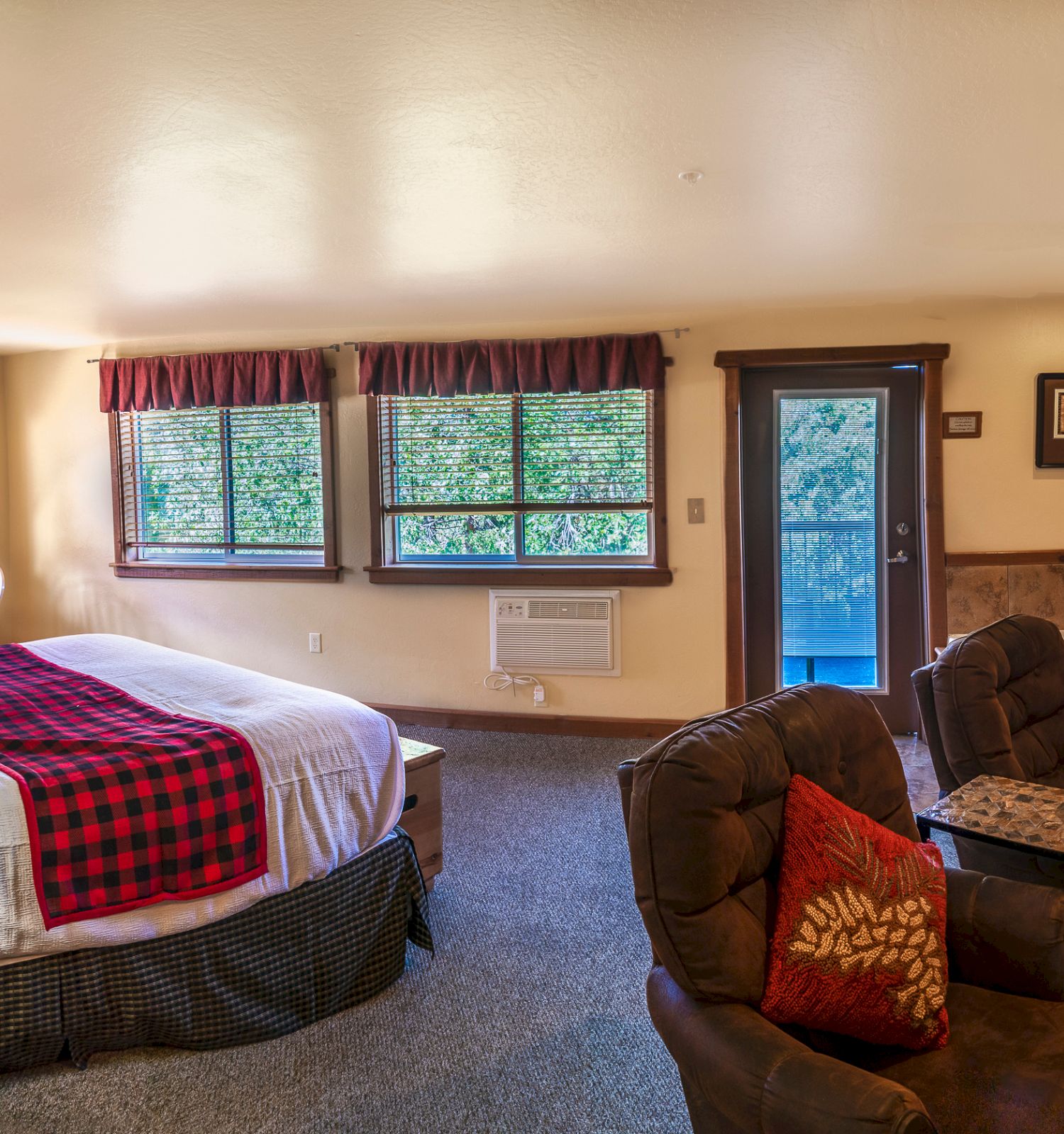 A cozy bedroom with a large bed, armchairs, a dresser, TV, and large windows. There is a small table, and an open door leads to a balcony.
