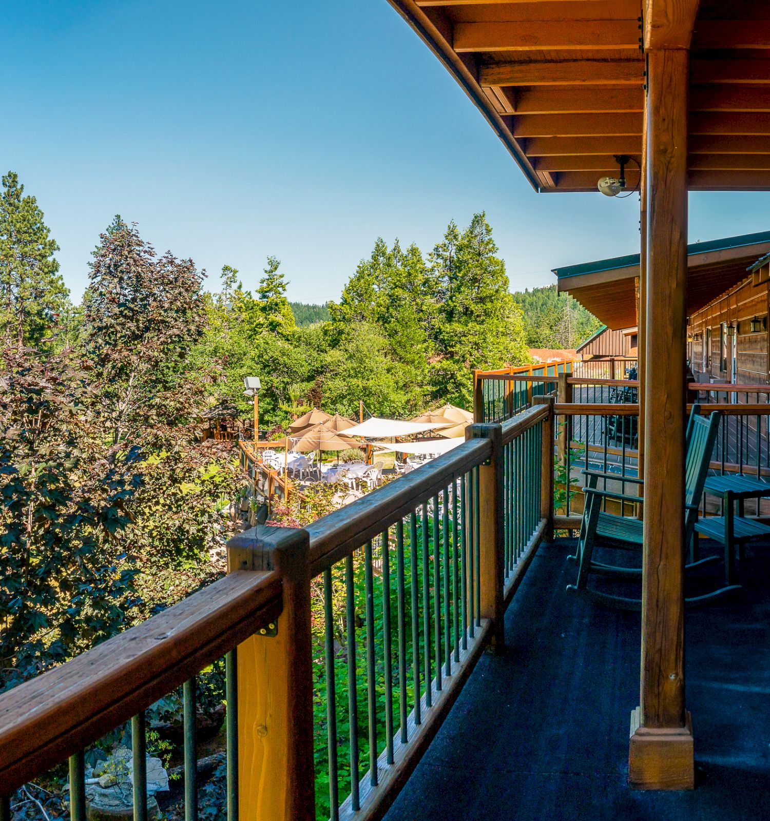 A wooden balcony with chairs overlooks a scenic forest view with clear blue skies, featuring a cozy and peaceful outdoor atmosphere.