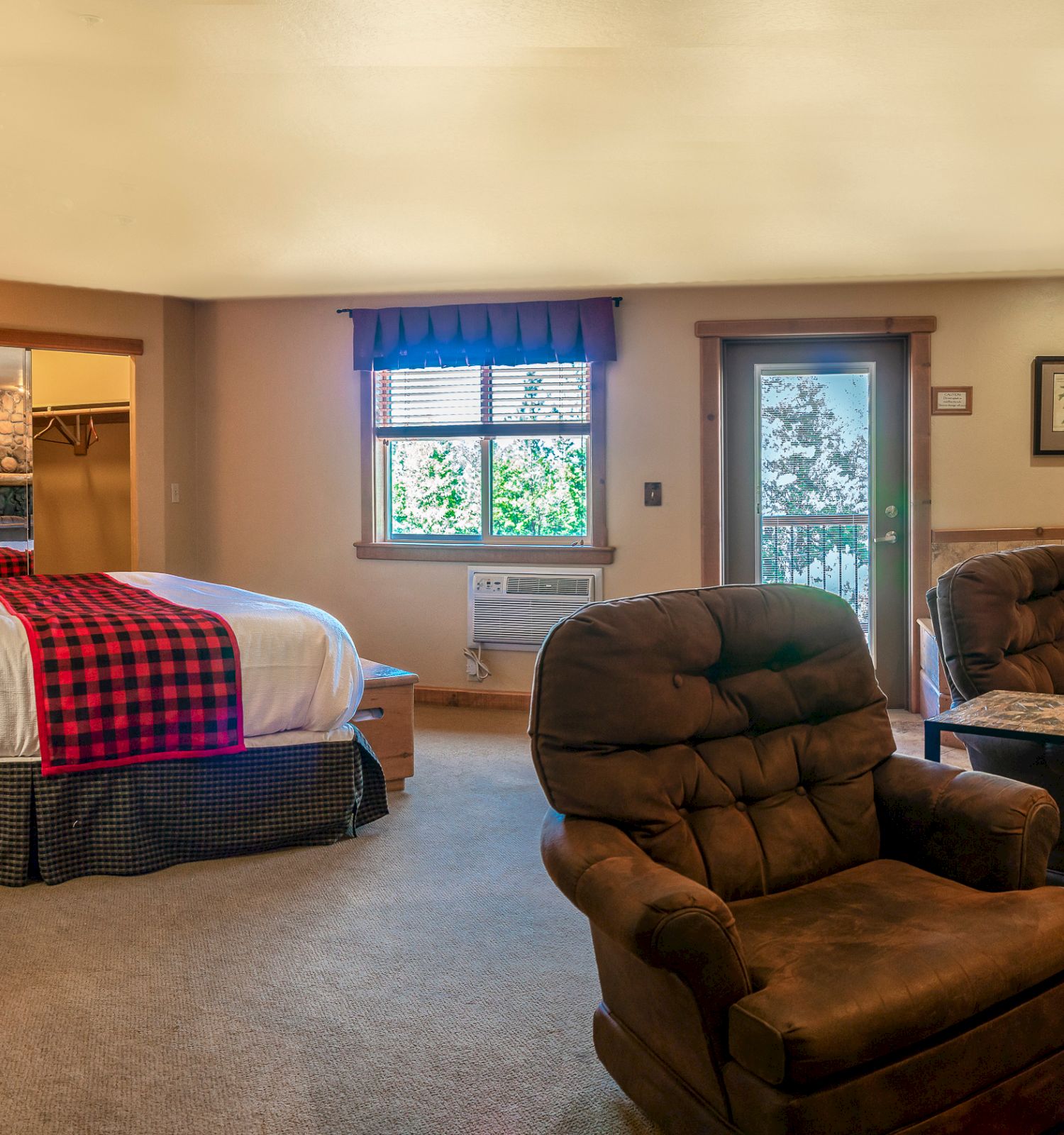 A cozy bedroom features a bed, two armchairs, a dresser, and a TV, with a door and window providing natural light.