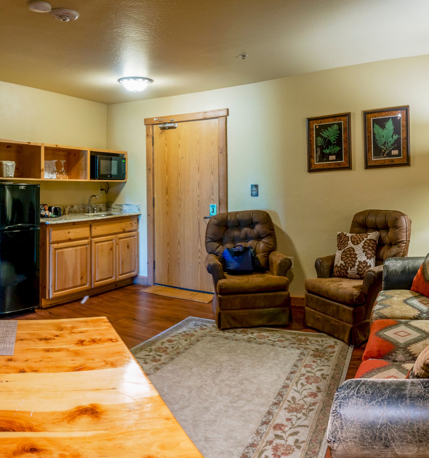 The image shows a cozy living room with a kitchen, a sofa, two armchairs, a dining table, a TV, a fridge, and framed pictures on the wall.