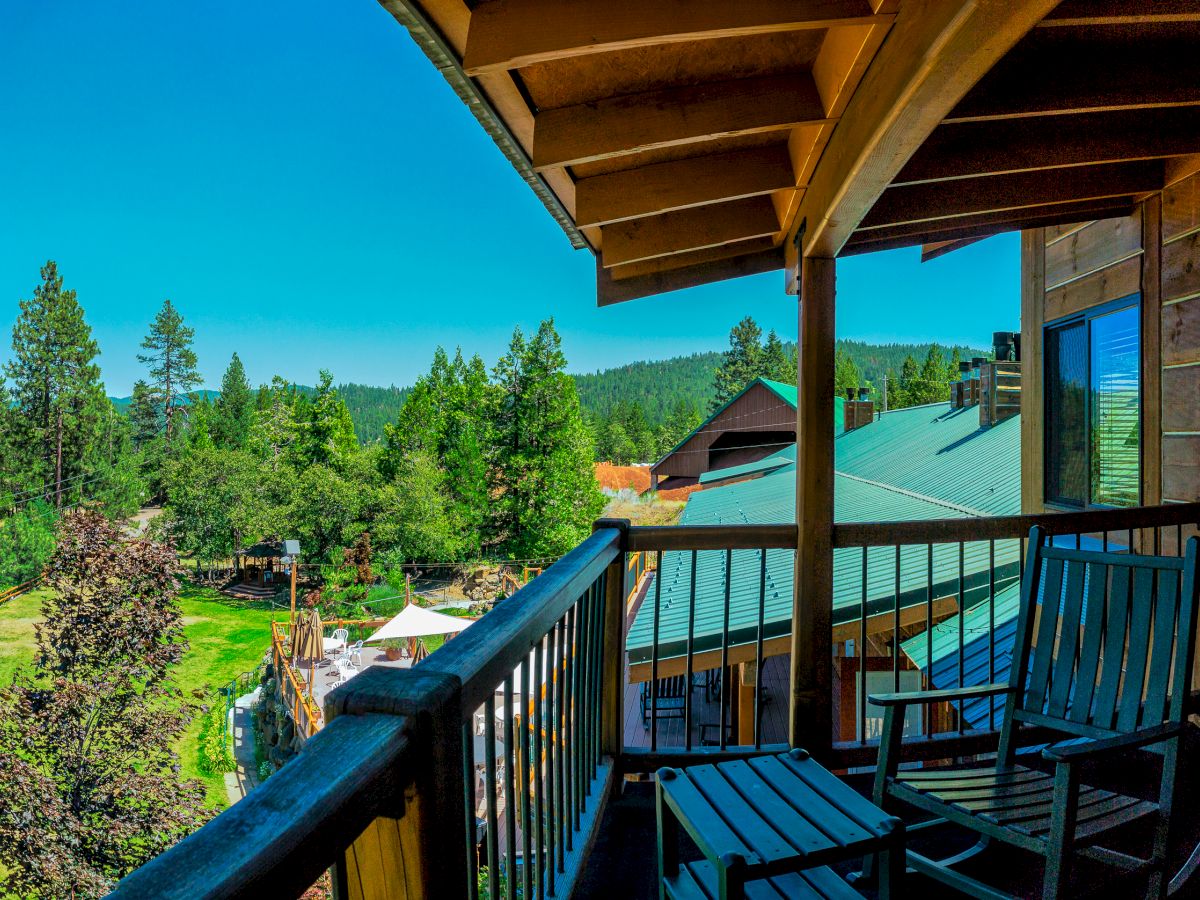 A scenic view from a wooden deck features two chairs overlooking a lush, forested area with clear blue skies, and rustic buildings in the background.