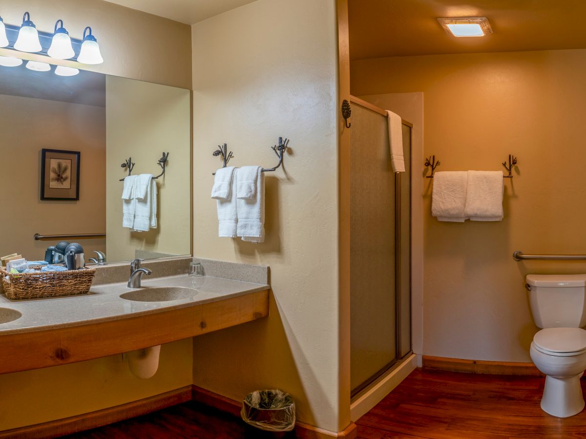 This image shows a bathroom with two sinks, a mirror, towels, a shower, a toilet, and wooden flooring in a warmly lit setting.