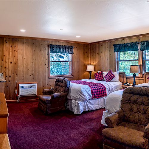 A cozy cabin-style bedroom with two beds, plaid bedding, wooden walls, armchairs, a TV on a wooden dresser, and side tables with lamps.