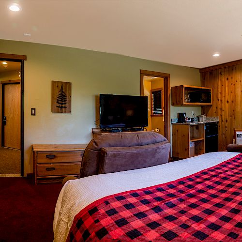 The image shows a cozy bedroom with two beds featuring red plaid blankets, a TV, a small table with chairs, and wood-paneled walls.
