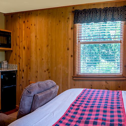 A cozy bedroom with wooden walls, a bed with red and black plaid accents, an armchair, a window with blinds, and a kitchenette area with a microwave.