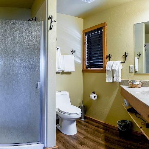 The image shows a clean bathroom with a walk-in shower, toilet, sink, mirror, towel racks, and wooden flooring. There is a window with blinds.